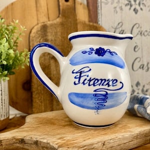 May include: White ceramic pitcher with blue accents and the word "Firenze" in script. The pitcher has a curved handle and a spout. Decorative blue floral and swirl designs are painted on the pitcher.