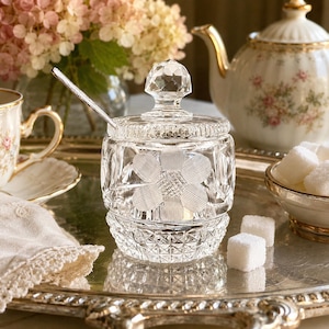May include: A clear, cut-glass sugar bowl with a faceted lid and a flower design. A small spoon rests inside. The bowl sits on a silver tray, alongside a teacup, teapot, and a small bowl of sugar cubes.