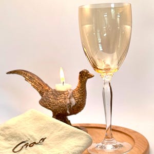May include: A decorative pheasant candle holder with a lit candle, a gold-tinted wine glass, and a napkin with "Ciao" embroidered on it, all arranged on a wooden tray. The candle holder and glass are the focal points.