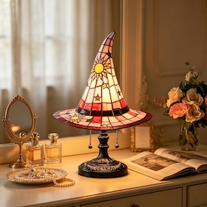 May include: A stained glass table lamp shaped like a pointed witch hat, featuring a white, red, and pink color palette with yellow sun and star motifs. The lamp has a dark metal base with a circular stand and two pull chains for operation.