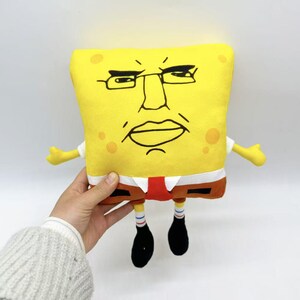 May include: A yellow plush toy resembling the cartoon character SpongeBob SquarePants, with an exaggerated facial expression. The toy has a white shirt, brown pants, and black shoes. The toy is being held by a person.