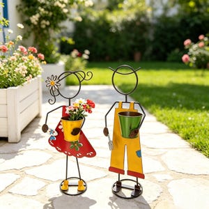 Könnte beinhalten: Zwei Metall-Gartenfiguren in Strichmännchen-Optik. Eine Figur trägt ein rotes Kleid mit weißen Punkten und hält einen gelben Eimer mit Blumen. Die andere Figur trägt eine gelbe Latzhose und hält einen grünen Eimer sowie eine kleine Metallgabel.