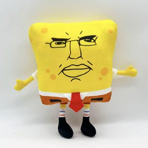 May include: A yellow plush toy resembling a cartoon character with a stern expression. The toy has a square body, a white shirt, a red tie, and brown shorts. It has black shoes and striped socks. The character's face features glasses and a frowning mouth.