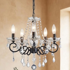 May include: Black wrought iron chandelier with crystal accents and five light bulbs. The chandelier hangs above a wooden dining table set with dishes and a vase of greenery. The word "ARTISANAL" is visible at the bottom.