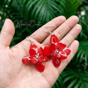 May include: A pair of red flower-shaped earrings with silver hooks. Each earring features a central pearl-like bead. The earrings are handmade and have a glossy finish. The image includes the text "Kitsune Craft Design Co."