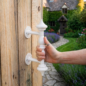 May include: White decorative gate handle with a detailed, ornate design. The handle is being held open, revealing a wooden gate and a pathway leading to a cottage. The handle is made of a white material.
