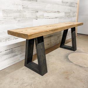 Puede incluir: Un banco rectangular de madera con un asiento grueso de madera de color marrón claro natural con veta visible y acabado liso. El banco se apoya sobre dos patas robustas en forma de trapecio, hechas de madera teñida oscura con veta vertical.