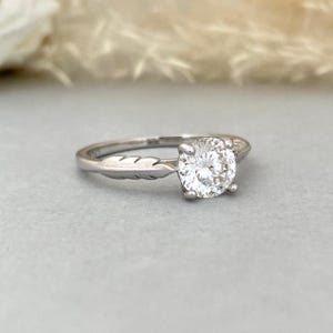 Op de afbeelding: Een zilveren ring met een grote, ronde, heldere edelsteen. De band heeft aan één kant een veermotief. De ring staat tegen een zachte grijze achtergrond, waardoor de schittering van de edelsteen wordt benadrukt.
