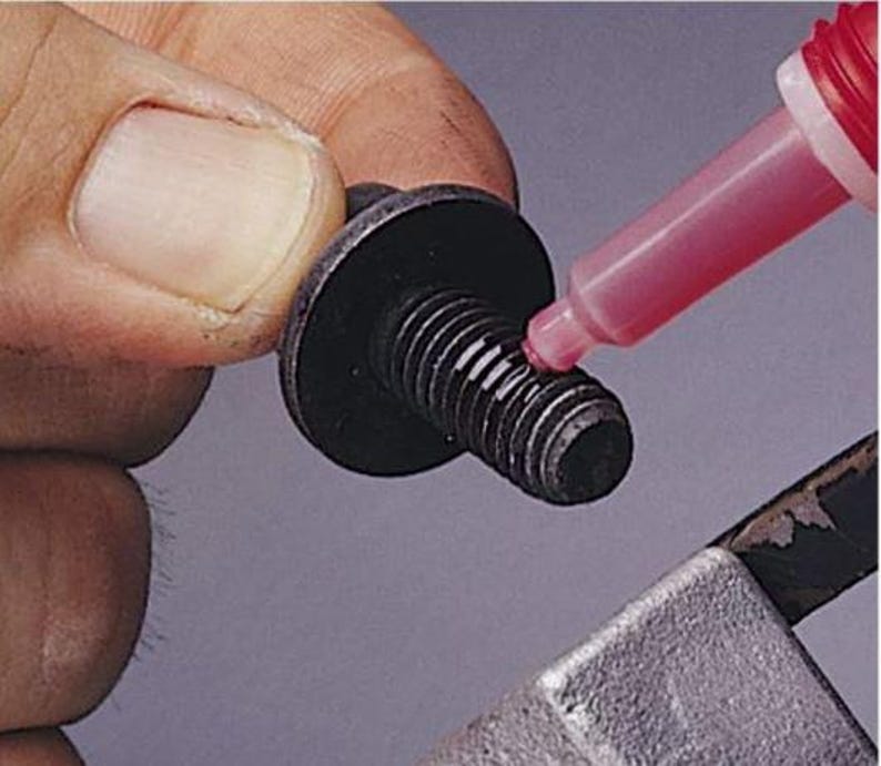 以下が含まれることがあります： 黒いボルトに、絞り出しボトルから赤い液体を塗布しているクローズアップ。ボルトは幅広で平らな頭部とねじ山のあるシャフトを持っています。赤い液体はねじ山に塗布されています。
