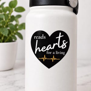 May include: A white water bottle with a black heart-shaped sticker. The sticker features the white text reads hearts for a living and a yellow EKG heartbeat line graphic at the bottom. The bottle has a black lid with a silver metallic accent.