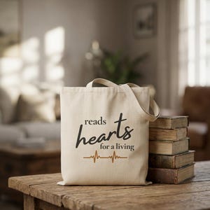 May include: A beige canvas tote bag featuring the black text reads hearts for a living and a brown heartbeat line graphic. The bag is positioned next to a stack of five vintage books with worn covers on a rustic wooden surface.