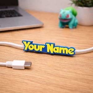 Può includere: Un organizer per cavi blu e giallo con la scritta "Your Name" su un cavo bianco. Il cavo è collegato a un connettore USB-C bianco. Un laptop e una figura verde di Bulbasaur sono sullo sfondo.