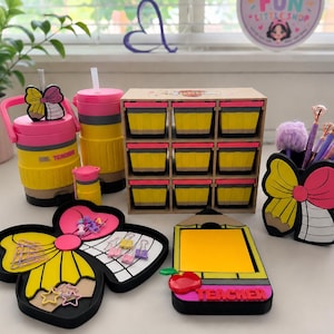 May include: A collection of teacher-themed desk accessories. Includes a pink and yellow insulated tumbler, a small storage organizer with yellow compartments, a pencil holder, and a notepad holder. The accessories are decorated with bows and the word "Teacher".