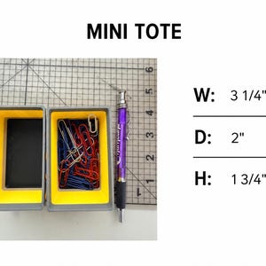 May include: A grey and yellow mini tote container filled with red, blue, and silver paper clips, positioned next to a purple mechanical pencil on a grid mat. Text reads MINI TOTE with dimensions W: 3 1/4", D: 2", and H: 1 3/4".