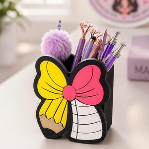 May include: A black desk organizer featuring a butterfly-shaped front panel with a yellow pencil design on the left wing and a white lined paper design on the right wing. The organizer holds several pens, including one with a purple pom-pom topper.