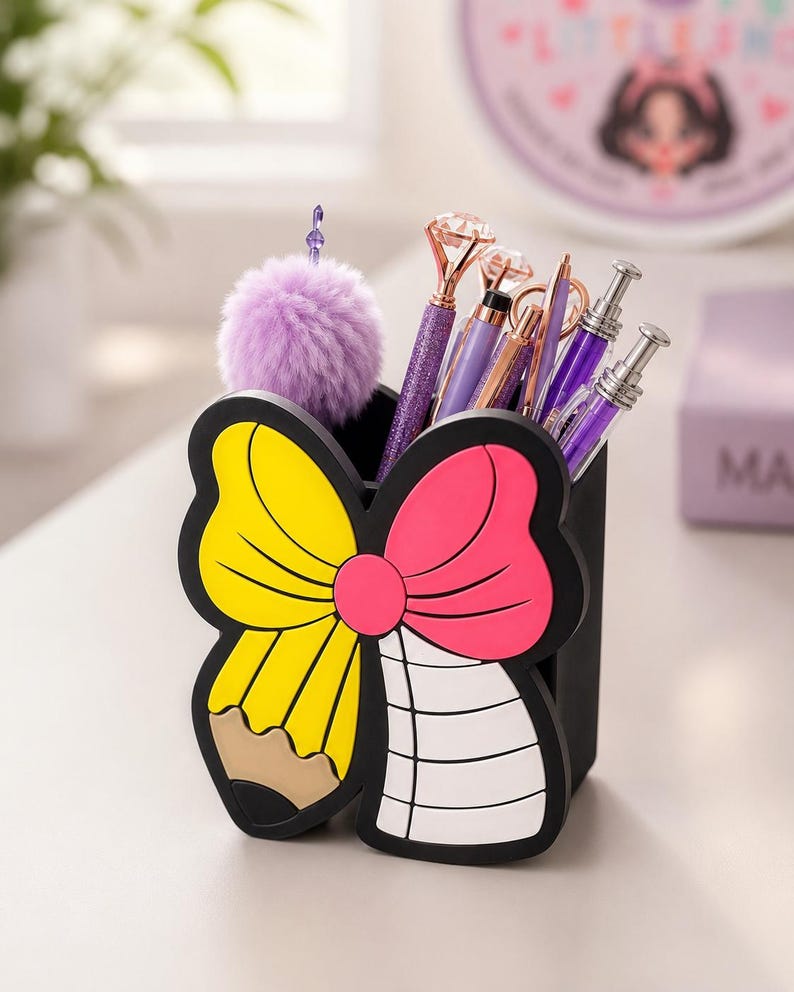 May include: A black desk organizer featuring a butterfly-shaped front panel with a yellow pencil design on the left wing and a white lined paper design on the right wing. The organizer holds several pens, including one with a purple pom-pom topper.