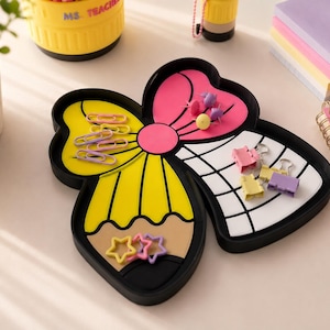 May include: A black desk organizer tray shaped like a bow, featuring a yellow pencil design on one side and a white grid pattern on the other. The tray holds various office supplies, including pink paper clips, star-shaped clips, and binder clips.
