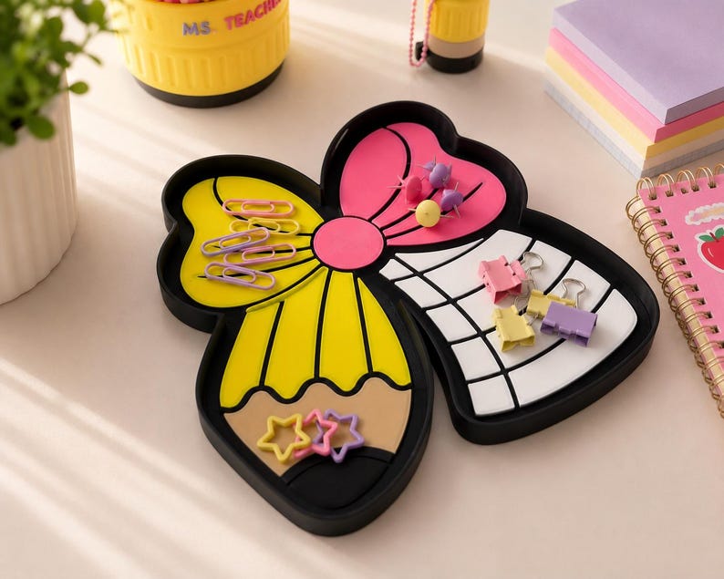 May include: A black desk organizer tray shaped like a bow, featuring a yellow pencil design on one side and a white grid pattern on the other. The tray holds various office supplies, including pink paper clips, star-shaped clips, and binder clips.