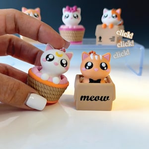 May include: Small collectible cat figurines with large eyes. One white cat with a yellow crescent moon sits in a woven basket. An orange cat with a black bow tie sits in a tan box labelled meow. Additional cat figures are visible in the background.
