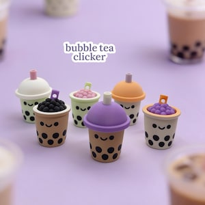 May include: A collection of six small bubble tea clickers featuring smiling faces and various coloured lids. Each miniature cup is decorated with black dots representing tapioca pearls, with some showing textured purple or black pearl tops.