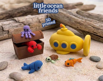 May include: A set of 3D printed little ocean friends figures including a brown treasure chest with a purple octopus and red lobster, a yellow submarine, a blue whale, a green sea turtle, and an orange fish on a sandy surface.