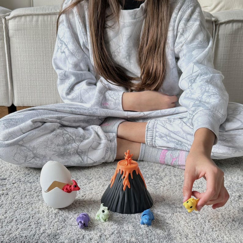 May include: A child's hand reaches for a small yellow dinosaur toy. The scene includes a black volcano toy with orange lava, a cracked white egg, and several colorful dinosaur figures. The toys are on a gray carpet.