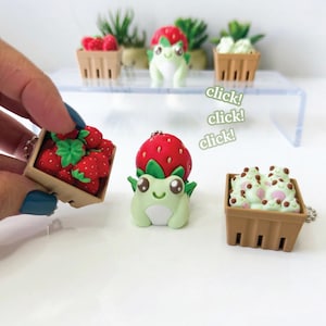 Könnte beinhalten: Drei Miniatur-Schlüsselanhänger mit hellgrünen Fröschen und roten Erdbeeren in braunen Körben. Ein Frosch trägt einen Erdbeerhut, ein Korb enthält rote Erdbeeren und ein Korb enthält kleine hellgrüne Frösche. Der Text lautet click! click! click!