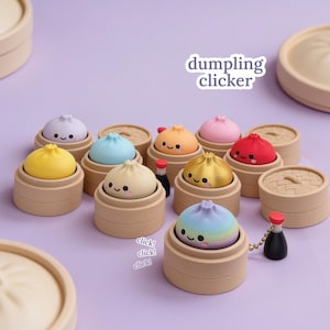 Puede incluir: Una colección de juguetes tipo clicker con forma de dumpling, cada uno con una cara sonriente y colocado en una pequeña vaporera de bambú beige. El set incluye varios colores sólidos, un dorado metálico y un diseño de arcoíris pastel.
