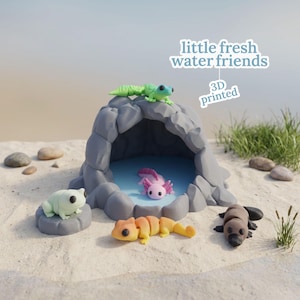 Puede incluir: Diorama impreso en 3D con una cueva gris con una piscina azul, rodeada de arena y pequeñas rocas. La escena incluye un gecko verde, un ajolote rosa, una rana verde claro, una salamandra naranja y amarilla y un ornitorrinco marrón. Texto: "little fresh water friends".