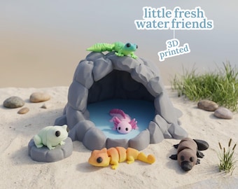 May include: A 3D-printed diorama featuring a gray cave with a blue pool, surrounded by sand and small rocks. The scene includes a green gecko, a pink axolotl, a light green frog, an orange and yellow salamander, and a brown platypus. Text reads "little fresh water friends".