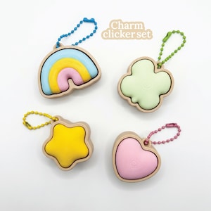 Könnte beinhalten: Ein Set aus vier Charm-Clickern in Form eines Regenbogens, eines vierblättrigen Kleeblatts, eines Sterns und eines Herzens. Jeder Anhänger hat eine farbige Mitte und einen beigen Rand, befestigt an einer Kette. Der Text "Charm clicker set" ist zu sehen.