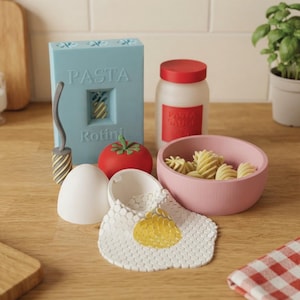 May include: A set of toy kitchen items including a light blue box labelled PASTA Rotini, a white jar with a red lid labelled PASTA SAUCE, a red tomato, a pink bowl filled with rotini pasta, a white egg, and a fried egg with a yellow yolk.