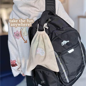 May include: A black backpack with a white beaver logo and a beige drawstring bag with the text "Little Printy Co." hanging from it. The image also shows a light gray hoodie with the text "take the fun anywhere!"