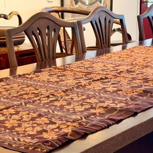 May include: A long, rectangular, dark brown table runner with a repeating floral pattern in shades of orange and gold. The runner is placed on a dark wooden table, with several wooden chairs visible in the background. The overall theme is home decor.
