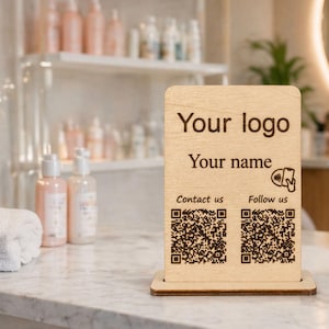 May include: A light-colored wooden sign with the text "Your logo" and "Your name" engraved on it. Below are the words "Contact us" and "Follow us" with corresponding QR codes. A small graphic is next to "Follow us". The sign is on a small wooden base.