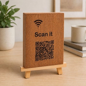 Puede incluir: Un bloque de madera con el texto "Scan it" y un código QR, exhibido en un pequeño caballete de madera. El bloque también presenta un símbolo de Wi-Fi. La madera tiene una veta natural y es de color marrón claro.
