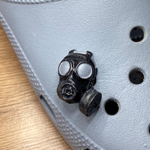 May include: A gray Crocs shoe with a black gas mask charm attached. The shoe has a red button on the side and multiple ventilation holes. The gas mask charm is detailed with silver accents.