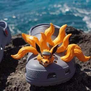 Puede incluir: Zuecos Crocs grises personalizados con un personaje naranja y negro con forma de zorro de nueve colas. El personaje es un diseño tridimensional detallado. Los zuecos están sobre una superficie rocosa con el océano al fondo.