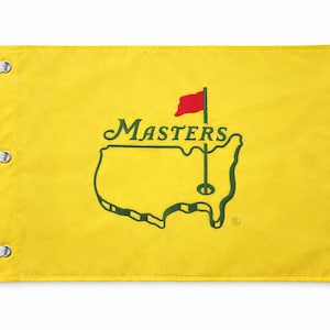 May include: Yellow flag featuring the Masters logo. The logo includes a green outline of Georgia, the word "Masters", and a red flag. The flag has three white grommets for hanging.