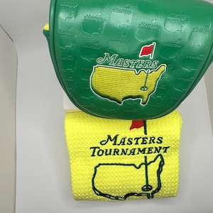 May include: A green golf club head cover and a yellow golf towel. The head cover features the Masters logo and is green with a textured pattern. The towel has the Masters Tournament logo and a map of the course.