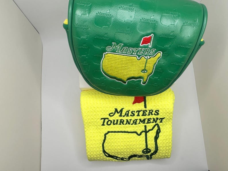 May include: A green golf club head cover and a yellow golf towel. The head cover features the Masters logo and is green with a textured pattern. The towel has the Masters Tournament logo and a map of the course.