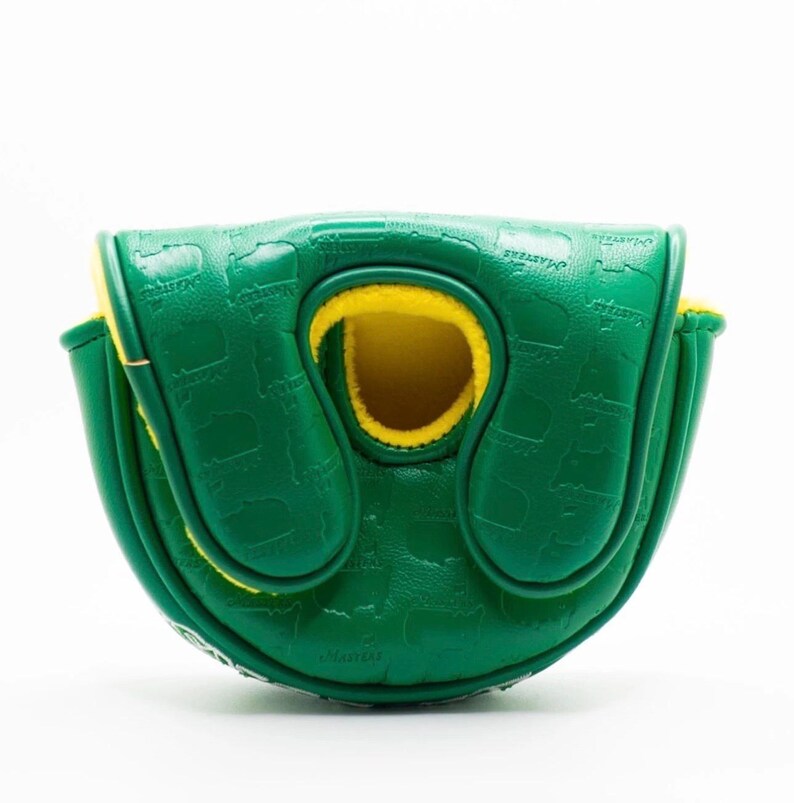 Puede incluir: Una funda verde para putter de golf con interior amarillo. La funda est&aacute; grabada con un patr&oacute;n repetitivo y la palabra "Masters". La funda est&aacute; hecha de un material duradero y tiene un cierre magn&eacute;tico.