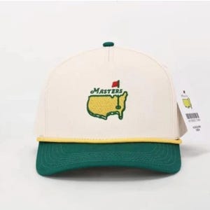 Può includere: Cappellino da baseball color crema con visiera verde e dettaglio corda gialla. Il cappellino presenta il logo Masters ricamato in verde e giallo, con la scritta "Masters" sopra. È attaccata una piccola etichetta.