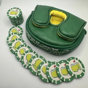 May include: A green golf putter cover and golf ball markers. The putter cover is green with the word "Masters" in white. The golf ball markers are white with a green border and a yellow map design with the word "Masters".