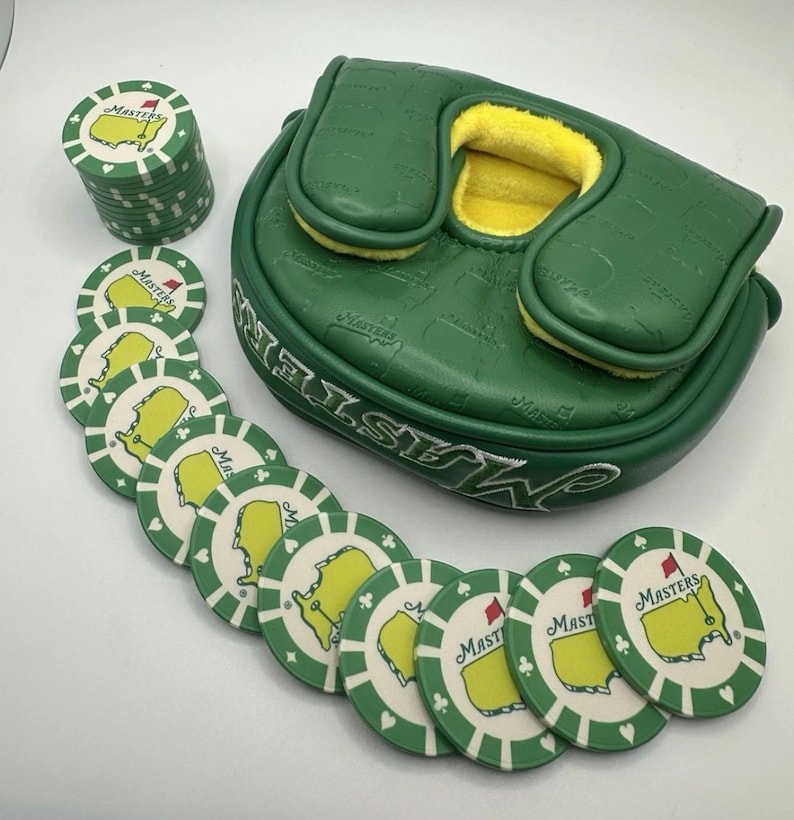 May include: A green golf putter cover and golf ball markers. The putter cover is green with the word "Masters" in white. The golf ball markers are white with a green border and a yellow map design with the word "Masters".