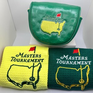 Puede incluir: Una funda verde para putter de golf y dos toallas de golf. La funda y las toallas presentan el logotipo del Masters Tournament, que incluye un mapa amarillo de los Estados Unidos con una bandera roja. Las toallas son amarillas y verdes.