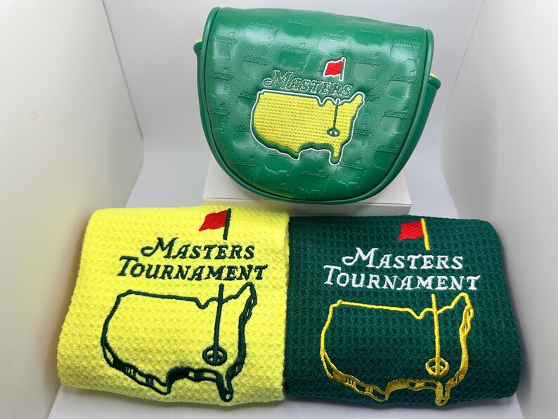 Puede incluir: Una funda verde para putter de golf y dos toallas de golf. La funda y las toallas presentan el logotipo del Masters Tournament, que incluye un mapa amarillo de los Estados Unidos con una bandera roja. Las toallas son amarillas y verdes.