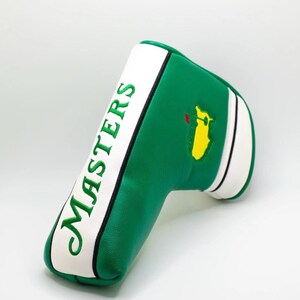 May include: Green and white golf club head cover with the word "MASTERS" embroidered in green. The cover features a yellow and green logo on the side. The cover is made of a smooth material.