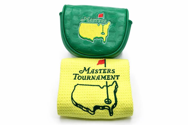 May include: Green golf club head cover and yellow golf towel. The head cover features the Masters logo and a red flag. The towel displays the Masters Tournament logo and a map of the course.