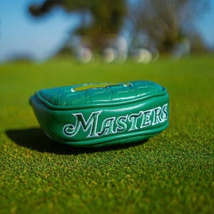 May include: A green golf club head cover with the word "Masters" embroidered in white. The cover is on a green golf course. The cover is made of a textured material.